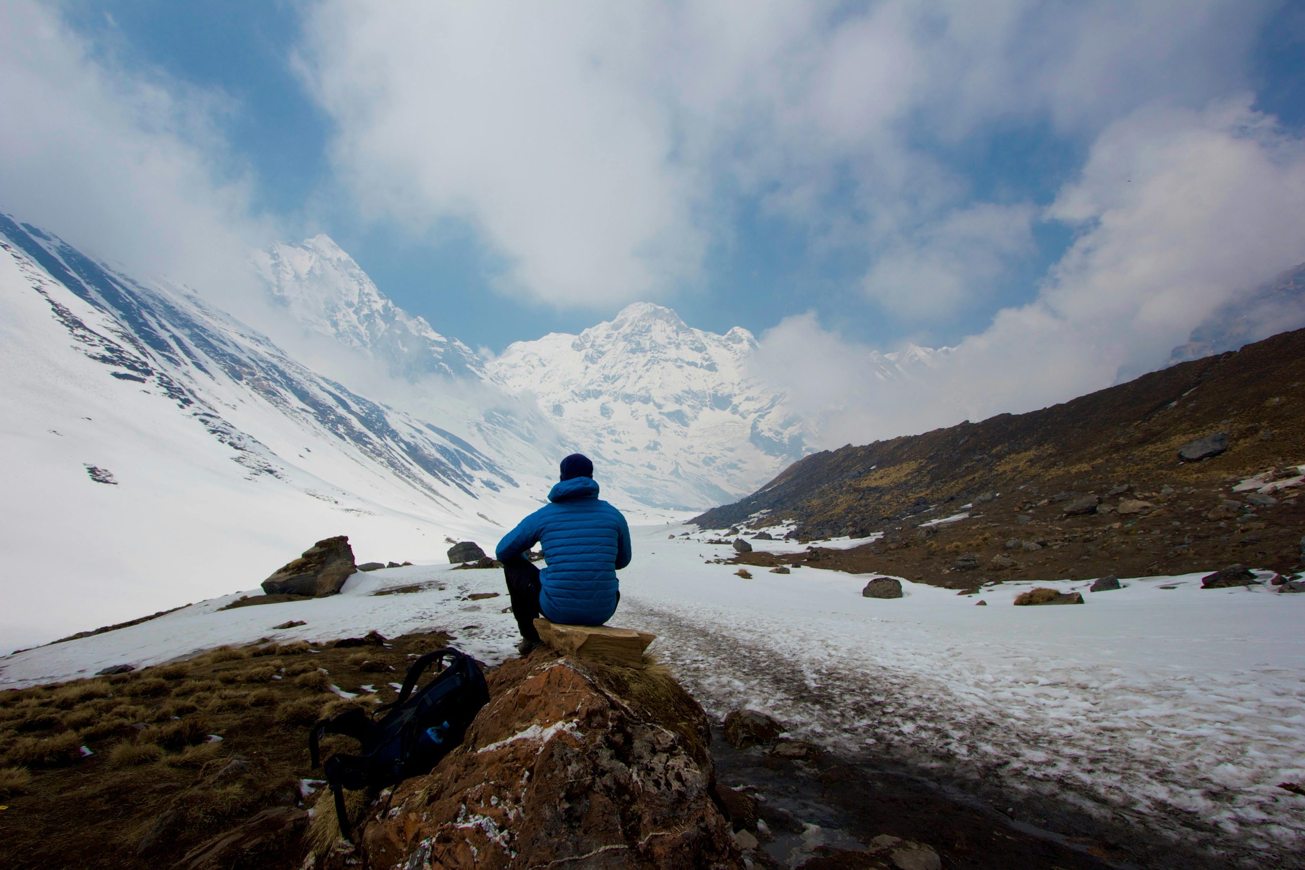 A man in a blue jacket sits in front of a snowy mountain range, embracing the tranquil scenery.